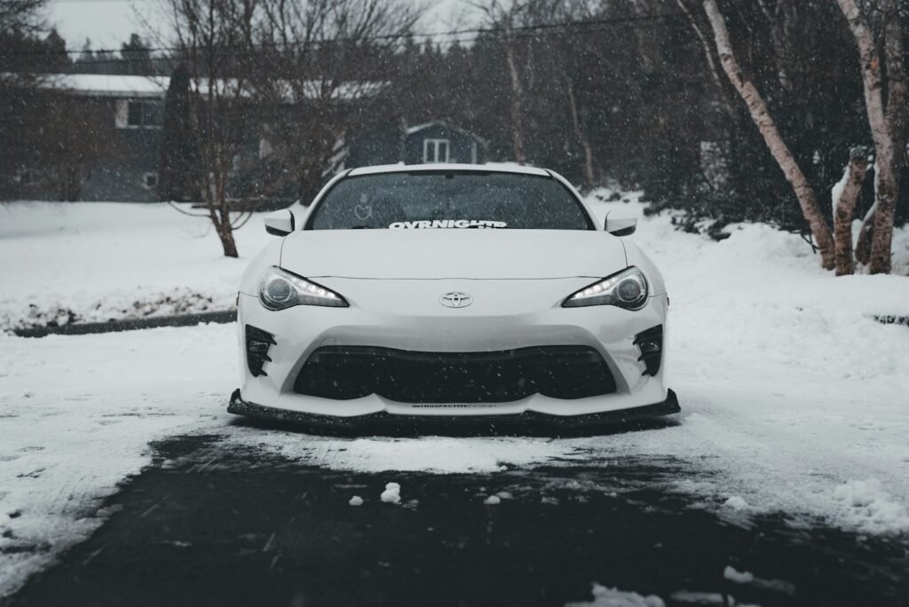 Carro branco estacionado em uma área com neve, cercado por árvores e casas ao fundo.