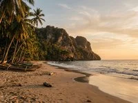 Praia de águas calmas com coqueiros, barcos pequenos e falésia ao fundo no fim da tarde