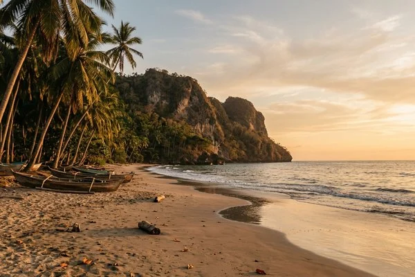 Praia de águas calmas com coqueiros, barcos pequenos e falésia ao fundo no fim da tarde