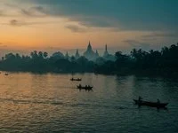 Rio largo ao entardecer com barcos pequenos e torres de templo ao fundo em luz dourada